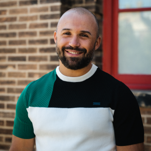 Indrit has light skin, a dark beard, and a shaved head. He wears a colour blocked tshirt and stands in front of a brick building smiling at the camera.