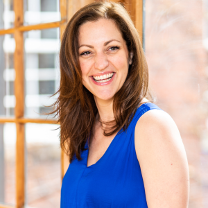 Kate has light skin and long brown hair, she wears a bright blue dress and leans against a window, smiling slightly away from the camera