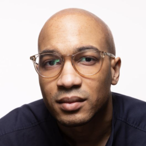 Chuck has medium toned skin, is bald, and wears tan coloured circular glasses. He wears a dark tshirt and looks into the camera.