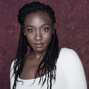 Virgilia is a Black woman, with long braided black hair. She wears a white shirt, stands against a maroon backdrop and looks into the camera