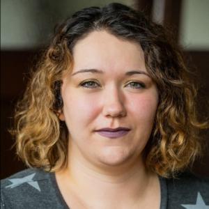 Caitlin has shoulder length curly hair with blonde highlights, and wears purple lipstick. 