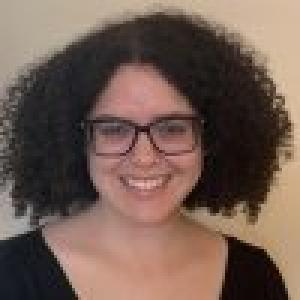 Signy has very curly dark hair cut into a bob, dark rimmed glasses, and is smiling at the camera