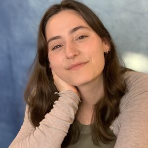 A women in a beige long sleeve rests her head on her chin while smiling at the camera in front of a blue background