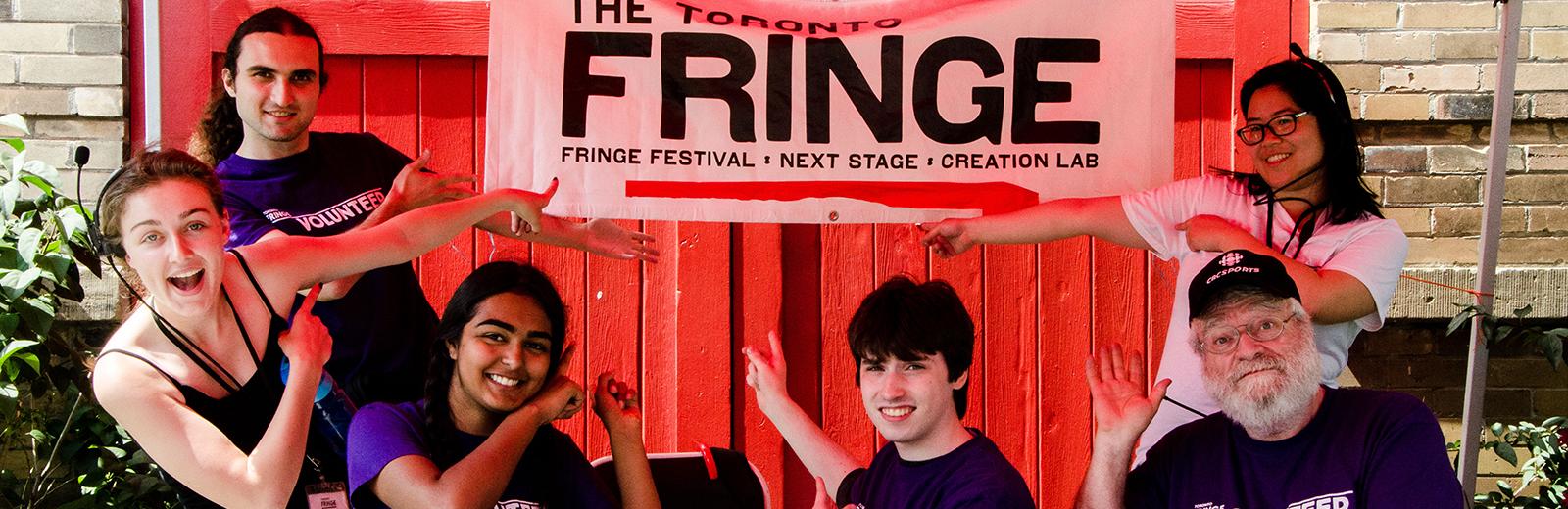 Staff and volunteers point to a Toronto Fringe sign, smiling at the camera