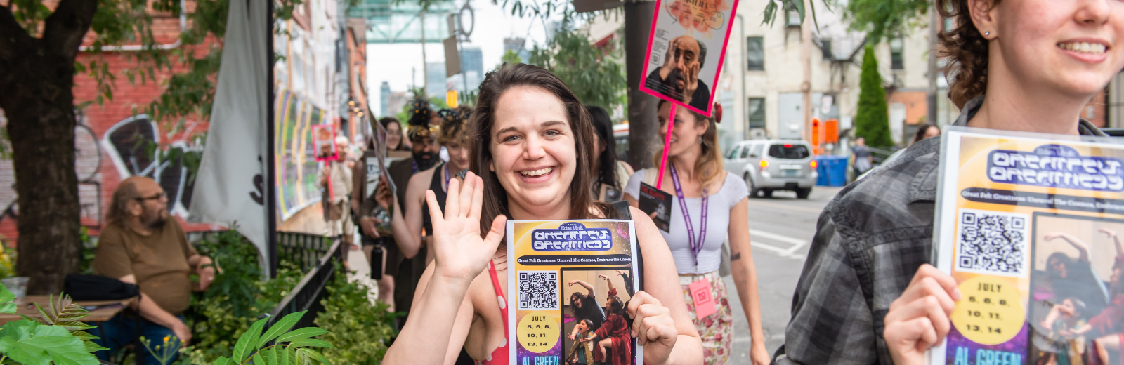 A Fringe artist smiles and waves while holding up their poster